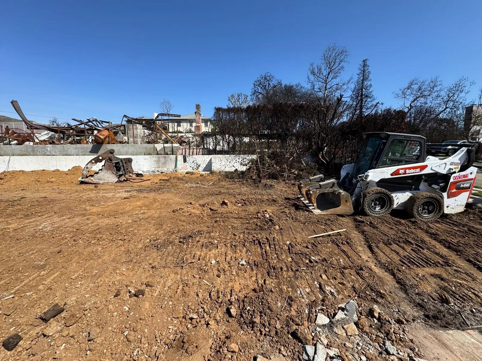 A Pacific Palisades lot cleared to bare soil after the fire, a skid-steer loader parked at the edge and the twisted remains of a neighboring structure still standing behind it.