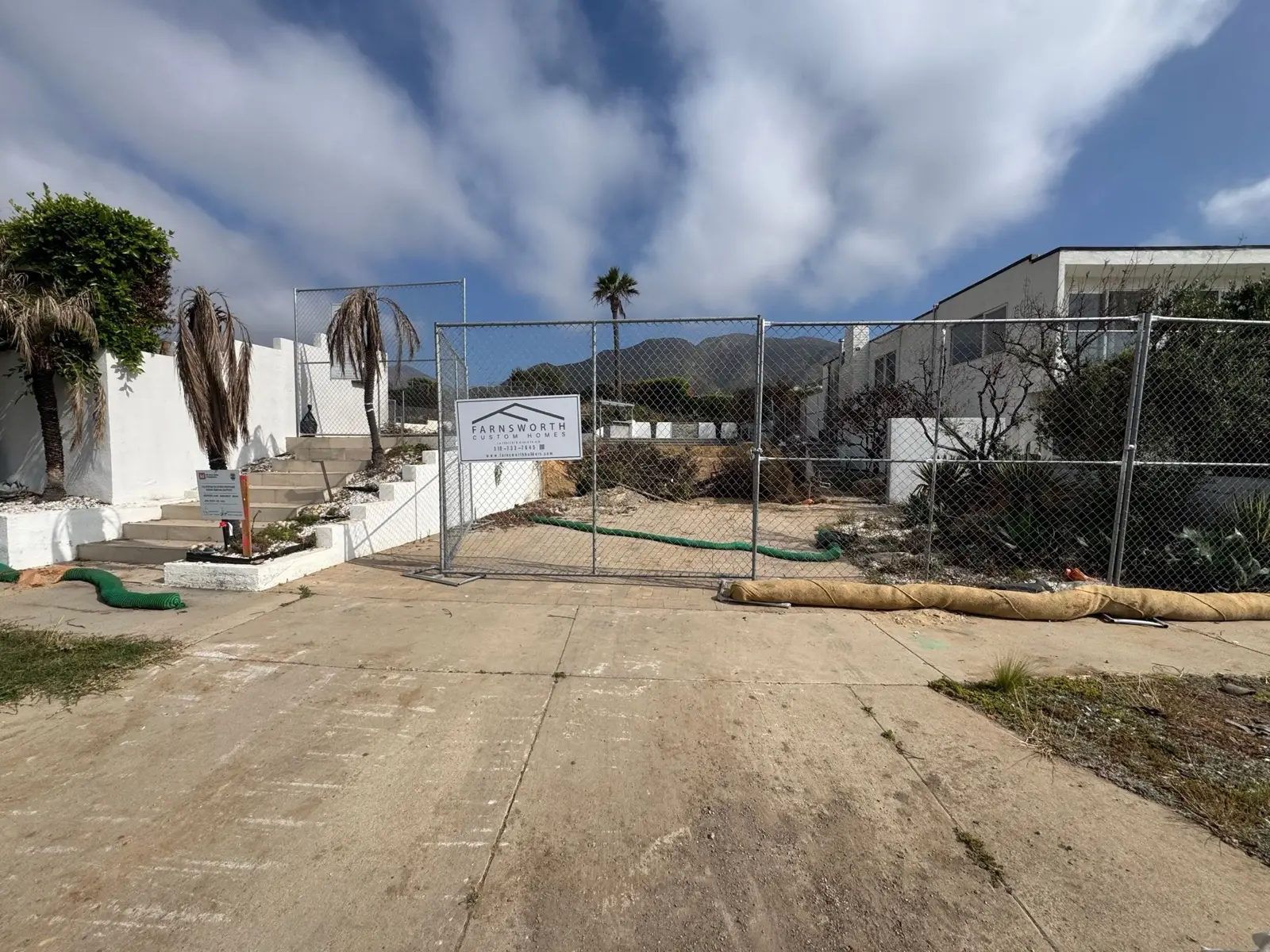 A cleared and fenced Pacific Palisades lot with a Farnsworth Builders sign on the perimeter fencing, mountains behind.